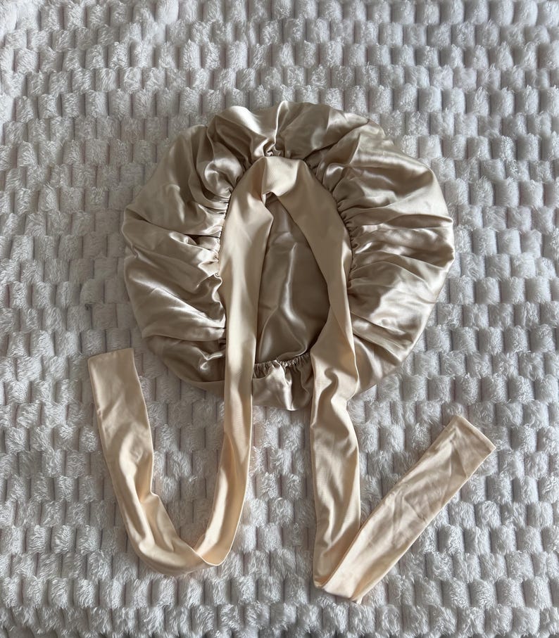 Pu&ograve; includere: Una cuffia per capelli in raso color champagne caratterizzata da un design circolare arricciato con una finitura liscia e lucente. La cuffia include due lunghi nastri in tessuto piatto che partono dal centro per una vestibilit&agrave; regolabile.