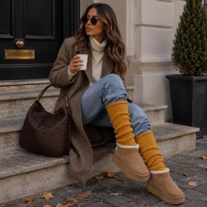 Op de afbeelding: Een bruine wollen jas, een witte coltrui, een lichtblauwe spijkerbroek, mosterdgele gebreide beenwarmers, beige suède platformlaarzen en een donkerbruine gevlochten leren tas. De outfit is gestyled met een zwarte zonnebril en een witte beker.