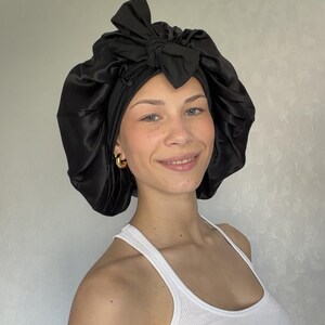 Pu&ograve; includere: Una cuffia per capelli in raso nero caratterizzata da un'ampia fascia arricciata e un fiocco decorativo annodato sul davanti. Il tessuto ha una finitura liscia e riflettente e il design include un bordo elastico per contenere i capelli.