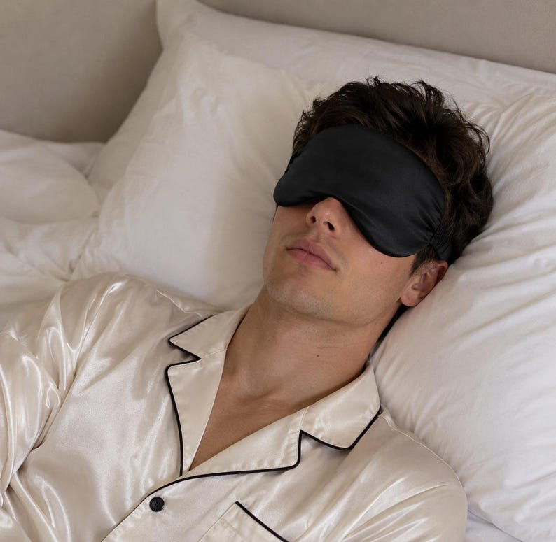 May include: A solid black sleep mask worn over the eyes, paired with a cream-coloured button-down pyjama top featuring a smooth, lustrous finish and thin black piping along the collar and front placket, worn while resting on a white pillow.