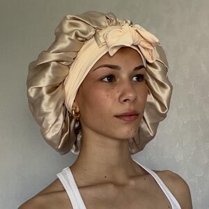 May include: A champagne-coloured satin sleep bonnet featuring a wide, pale peach-coloured elasticised headband with a decorative bow detail at the front. The bonnet has a gathered, voluminous design with a smooth, shiny finish and a soft fabric texture.