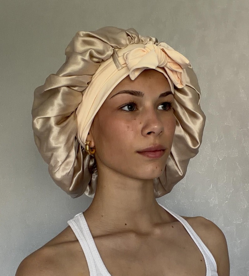 May include: A champagne-coloured satin sleep bonnet featuring a wide, pale peach-coloured elasticised headband with a decorative bow detail at the front. The bonnet has a gathered, voluminous design with a smooth, shiny finish and a soft fabric texture.