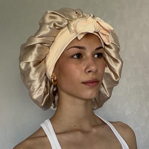 Pu&ograve; includere: Una cuffia da notte in raso color champagne con un'ampia fascia elastica color pesca pallido e un fiocco decorativo sul davanti. La cuffia presenta un design arricciato e voluminoso con una finitura liscia, lucida e una consistenza morbida.