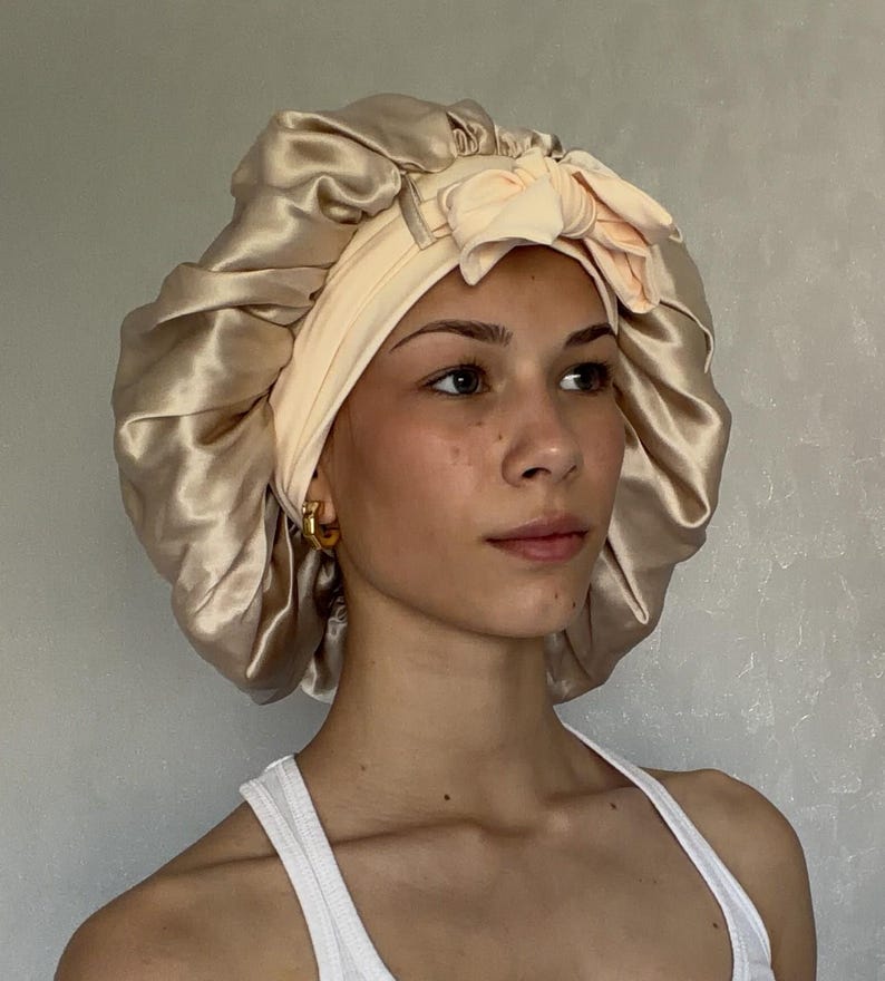 Pu&ograve; includere: Una cuffia da notte in raso color champagne con un'ampia fascia elastica color pesca pallido e un fiocco decorativo sul davanti. La cuffia presenta un design arricciato e voluminoso con una finitura liscia, lucida e una consistenza morbida.