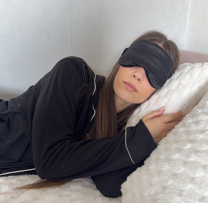 May include: A black sleep mask covers the eyes of a person lying on a white fluffy pillow. The person is wearing a black pajama set with white trim. The scene suggests relaxation and sleep.