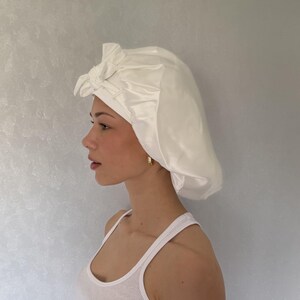 Pu&ograve; includere: Una cuffia per capelli in raso bianco caratterizzata da un design arricciato e da un dettaglio a nodo decorativo sulla parte anteriore. La cuffia ha una finitura liscia e lucente ed &egrave; realizzata con una fascia larga che aderisce alla fronte.
