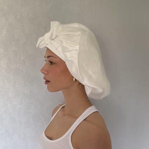 May include: A white satin hair bonnet featuring a gathered design and a decorative knotted bow detail at the front. The bonnet has a smooth, lustrous finish and is constructed with a wide band that sits securely around the forehead area.