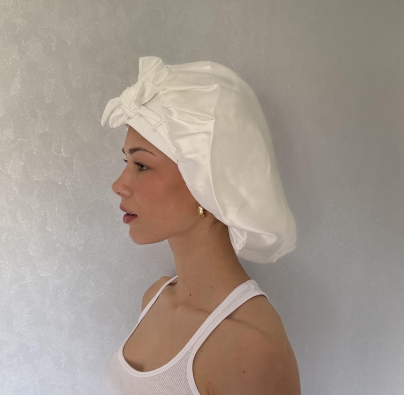 May include: A white satin hair bonnet featuring a gathered design and a decorative knotted bow detail at the front. The bonnet has a smooth, lustrous finish and is constructed with a wide band that sits securely around the forehead area.