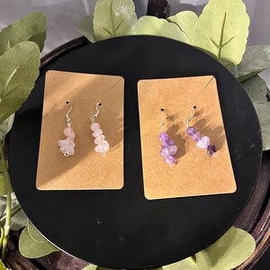 May include: Two pairs of earrings with silver hooks and multiple gemstone beads. One pair features pink beads and the other pair features purple beads.