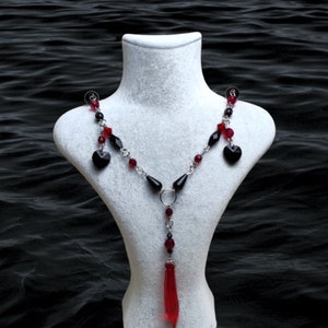 May include: A silver chain necklace with red and black crystal beads and a red crystal pendant. The necklace features two black heart-shaped charms.