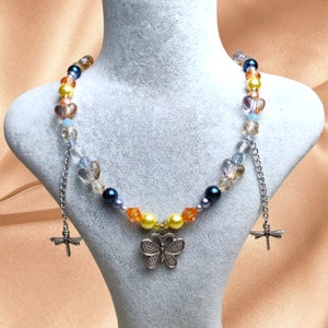 Può includere: Una collana di perline con un ciondolo a farfalla. La collana presenta un mix di perline colorate, tra cui perline gialle, arancioni, blu e trasparenti. Ciondoli a forma di libellula argentati pendono dalla catena. La collana è esposta su un manichino.