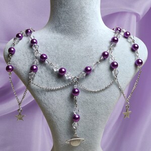 May include: A purple and silver body chain necklace with a celestial theme. The necklace features purple faux pearls, clear crystal beads, and silver chain accents. It includes a planet charm and star charms.