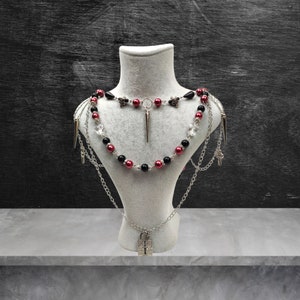 May include: A layered necklace with black and burgundy beads, silver chains, and a silver padlock pendant. The necklace is displayed on a white mannequin bust.