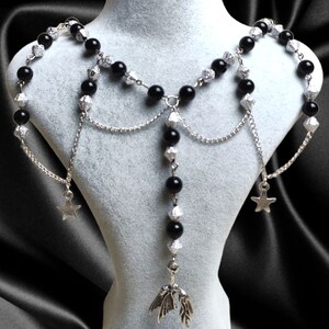 May include: A silver shoulder necklace with black beads and star charms. The necklace has multiple strands of delicate chains and decorative silver elements. The design includes a central pendant with a leaf-like shape.