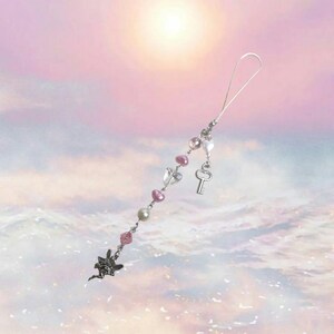 May include: A silver charm with a fairy design, pink and white beads, a silver key charm, and a silver chain. The charm is hanging from a silver string.