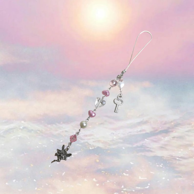 May include: A silver charm with a fairy design, pink and white beads, a silver key charm, and a silver chain. The charm is hanging from a silver string.