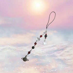 May include: A silver charm with a planet, star, and hearts on a black string. The charm is hanging from a black phone strap.
