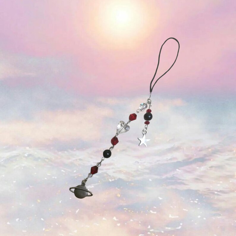 May include: A silver charm with a planet, star, and hearts on a black string. The charm is hanging from a black phone strap.