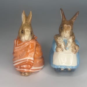 Może przedstawiać: Dwie ceramiczne figurki królika. Jeden królik jest owinięty pomarańczowym kocem z białymi paskami. Drugi królik ma na sobie niebieską sukienkę i biały fartuch. Oba mają brązowe uszy i twarze.