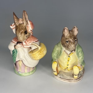 May include: Two ceramic figurines of anthropomorphic animals. One is a rabbit wearing a bonnet and holding a basket. The other is a mouse in a green jacket and yellow vest. Both are painted with detailed features and pastel colours.