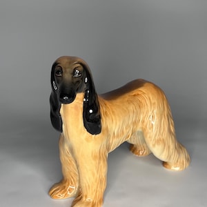 Puede incluir: Figura de cerámica de un galgo afgano. El perro está representado de pie con un pelaje largo y fluido. La figura presenta una paleta de colores beige, negro y marrón, con un acabado brillante. La cabeza del perro está detallada con orejas negras y un hocico oscuro.