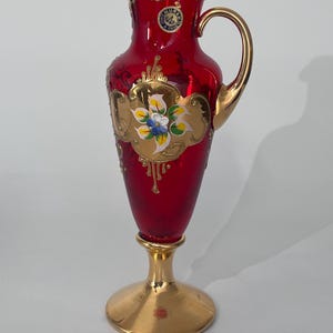 Könnte beinhalten: Eine rote Glaskaraffe mit goldenem Griff und Fuß. Die Karaffe hat goldene Verzierungen und ein Blumendesign mit blauen, gelben und weißen Akzenten. Ein kleiner runder Aufkleber ist oben sichtbar.