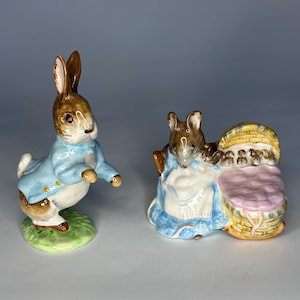 Puede incluir: Dos figuras de cerámica: un conejo marrón con una chaqueta azul claro y un ratón con un cochecito. El conejo está sobre una base verde, mientras que el ratón está junto a un cochecito amarillo y rosa. Las figuras son detalladas y coloridas.