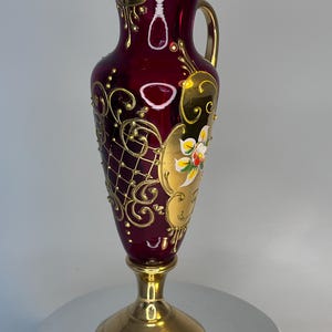 May include: A deep red glass pitcher featuring an ornate gold painted lattice pattern and floral motifs. The vessel has a gold-coloured handle, a flared rim, and sits on a polished gold-toned pedestal base, creating a tall and elegant silhouette.