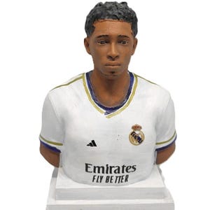Puede incluir: Una figura de busto en blanco y dorado de un jugador de fútbol que lleva una camiseta blanca con ribetes azules y dorados. La camiseta tiene el logotipo de Adidas en el lado derecho y el escudo del Real Madrid en el lado izquierdo. La camiseta también tiene el texto "Emirates Fly Better" impreso en la parte delantera.