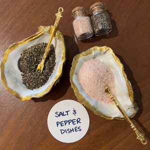 May include: Two gold oyster shell salt and pepper dishes with matching gold spoons. The dishes are filled with pink Himalayan salt and black peppercorns. A white circle label reads "SALT & PEPPER DISHES".
