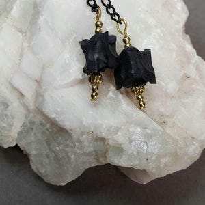 May include: A pair of black dangle earrings with gold accents. The earrings feature a black, square-shaped charm with a textured surface. The charms are attached to a black chain with gold beads.