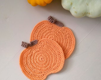 May include: Two crocheted orange pumpkin-shaped coasters with brown stems. The coasters are on a white surface.
