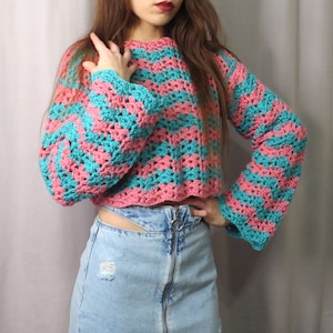 Könnte beinhalten: Ein gestrickter Pullover mit rosa und türkisfarbenen Streifen und Glockenärmeln. Der Pullover ist kurz und hat eine lockere Passform.