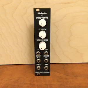 May include: Black synthesizer module with white knobs labeled "Frequency", "Spread", and "Harmonics". It has jacks labeled "IV", "SPRD CV", "FM", "HARM CV", "SINE", and "SQU". The module is labeled "SideBander" and "MIDIVERSE MODULAR".