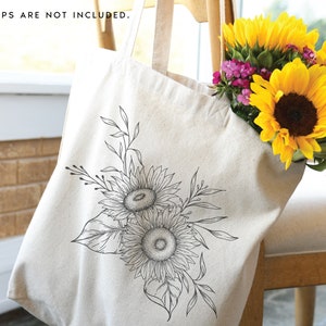 K&ouml;nnte beinhalten: Eine wei&szlig;e Canvas-Tragetasche mit einer schwarzen Linienzeichnung von Sonnenblumen und Bl&auml;ttern. Die Tasche liegt auf einem Holzstuhl mit einem Strau&szlig; gelber Sonnenblumen im Hintergrund.