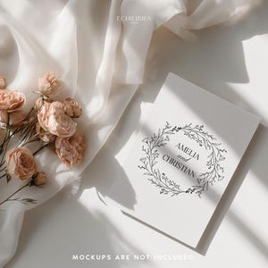 May include: A flat lay featuring a white invitation with the names "Amelia and Christian" inside a floral wreath. Peach-colored roses and a white fabric are arranged on a white surface. The text "MOCKUPS ARE NOT INCLUDED" is at the bottom.