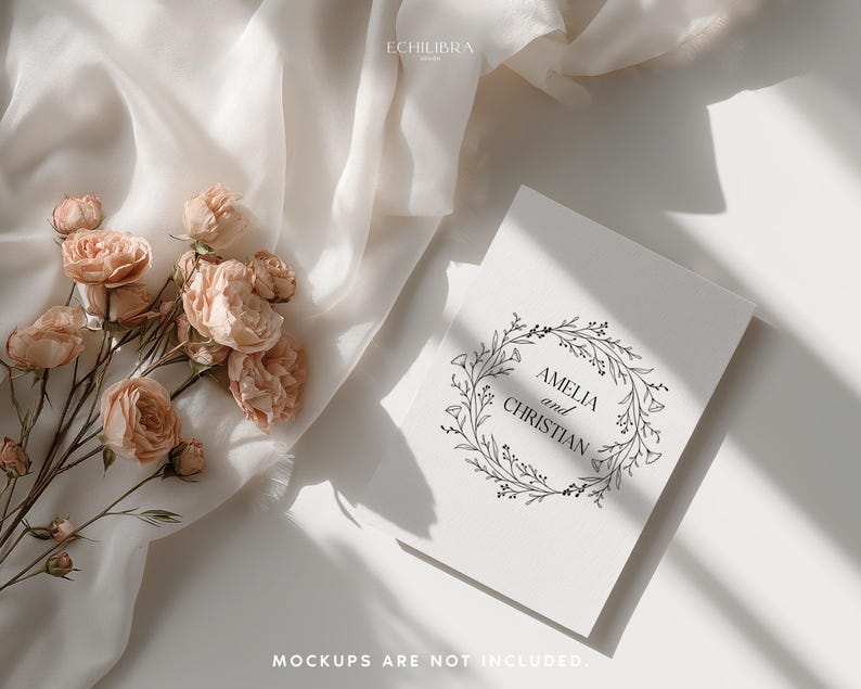 May include: A flat lay featuring a white invitation with the names "Amelia and Christian" inside a floral wreath. Peach-colored roses and a white fabric are arranged on a white surface. The text "MOCKUPS ARE NOT INCLUDED" is at the bottom.