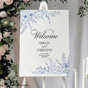 May include: A rectangular wedding welcome sign on a white easel featuring blue floral line art in the top left and bottom right corners. The text reads Welcome, AMELIA and CHRISTIAN, June 15, 2024, Rosewood Gardens, Springfield in black font.