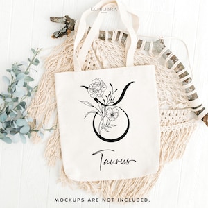 Puede incluir: Una bolsa de tela blanca con un dibujo de l&iacute;nea negra del signo del zod&iacute;aco Tauro adornado con flores. El texto "Taurus" est&aacute; escrito debajo del signo.