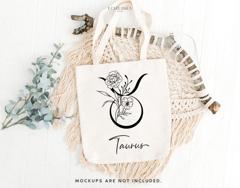 Puede incluir: Una bolsa de tela blanca con un dibujo de l&iacute;nea negra del signo del zod&iacute;aco Tauro adornado con flores. El texto "Taurus" est&aacute; escrito debajo del signo.