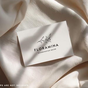 Puede incluir: Una tarjeta de visita rectangular blanca con un dibujo lineal negro de una rama con hojas sobre el texto FLORAMINA y el texto m&aacute;s peque&ntilde;o SCANDINAVIAN DECOR. La tarjeta est&aacute; colocada sobre una tela suave de color blanco roto.