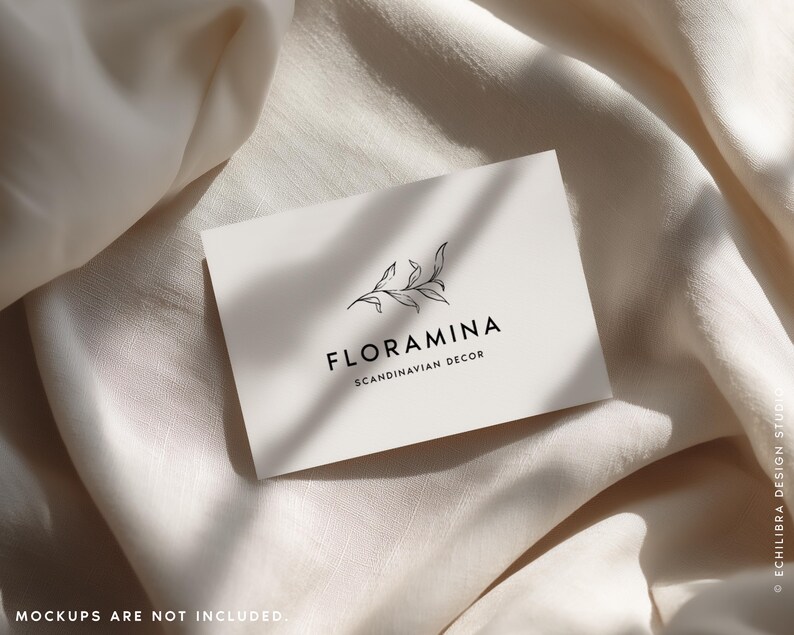 Puede incluir: Una tarjeta de visita rectangular blanca con un dibujo lineal negro de una rama con hojas sobre el texto FLORAMINA y el texto m&aacute;s peque&ntilde;o SCANDINAVIAN DECOR. La tarjeta est&aacute; colocada sobre una tela suave de color blanco roto.