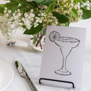 May include: A black and white illustration of a margarita glass with a lime wedge on a white card. The card is standing on a black metal stand. The card is on a white table with a white tablecloth.