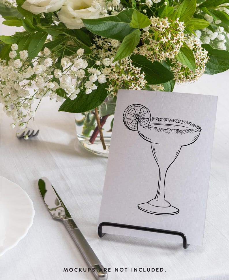 May include: A black and white illustration of a margarita glass with a lime wedge on a white card. The card is standing on a black metal stand. The card is on a white table with a white tablecloth.