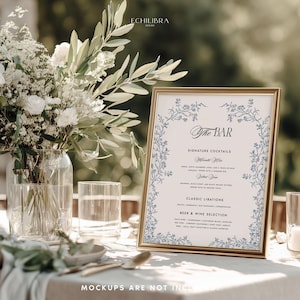 May include: A gold-framed bar menu sign featuring blue floral illustrations, black text reading The Bar, Signature Cocktails, Classic Libations, and Beer & Wine Selection, displayed next to a glass vase of white flowers and greenery.