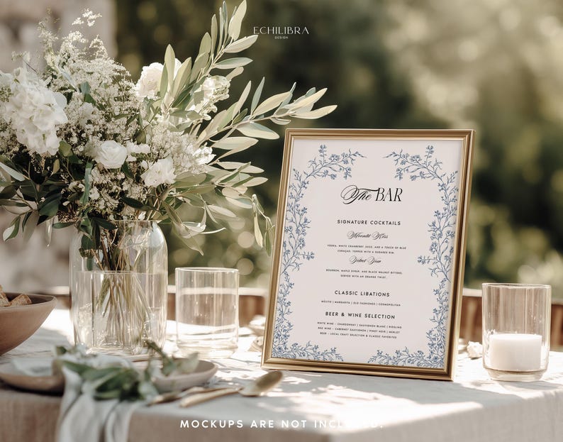 May include: A gold-framed bar menu sign featuring blue floral illustrations, black text reading The Bar, Signature Cocktails, Classic Libations, and Beer & Wine Selection, displayed next to a glass vase of white flowers and greenery.