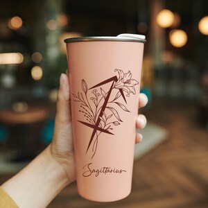 Puede incluir: Un vaso de acero inoxidable rosa con un dise&ntilde;o floral y el signo del zod&iacute;aco Sagitario. El vaso tiene una tapa negra y un interior blanco.