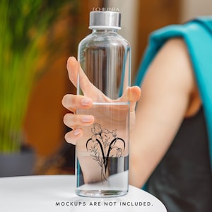 Puede incluir: Una botella de agua de vidrio transparente con un dise&ntilde;o floral negro en el lateral. La botella est&aacute; siendo sostenida por la mano de una persona.
