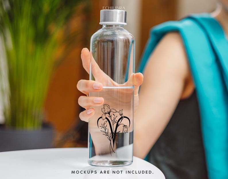 Puede incluir: Una botella de agua de vidrio transparente con un dise&ntilde;o floral negro en el lateral. La botella est&aacute; siendo sostenida por la mano de una persona.
