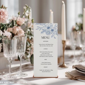 May include: A vertical rectangular menu card featuring a blue floral line art illustration at the top, the word MENU in black, and a list of food items in black text. The card is held upright by a thin metallic base on a white table setting.